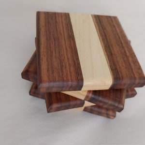 May include: A stack of four square wooden coasters. Each coaster is made of three vertical strips of wood: dark brown, light wood, and dark brown. The coasters are stacked at a slight angle, revealing their layered construction.