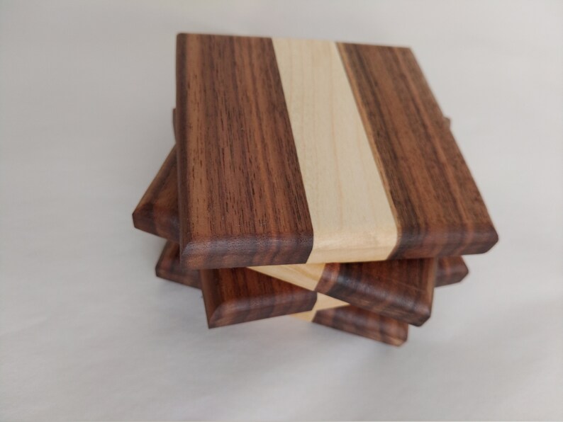 May include: A stack of four square wooden coasters. Each coaster is made of three vertical strips of wood: dark brown, light wood, and dark brown. The coasters are stacked at a slight angle, revealing their layered construction.