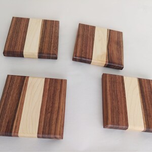 May include: Four square wooden coasters. Each coaster is made of dark brown wood with a light brown stripe down the center. The coasters are arranged on a white surface.