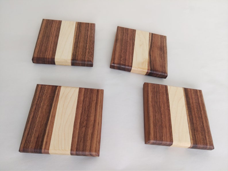 May include: Four square wooden coasters. Each coaster is made of dark brown wood with a light brown stripe down the center. The coasters are arranged on a white surface.