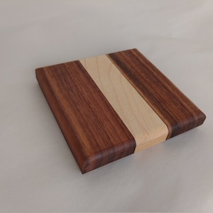 May include: Square wooden cutting board with a dark brown walnut exterior and a light maple wood stripe down the center. The board has rounded edges and a smooth finish, suitable for kitchen use.