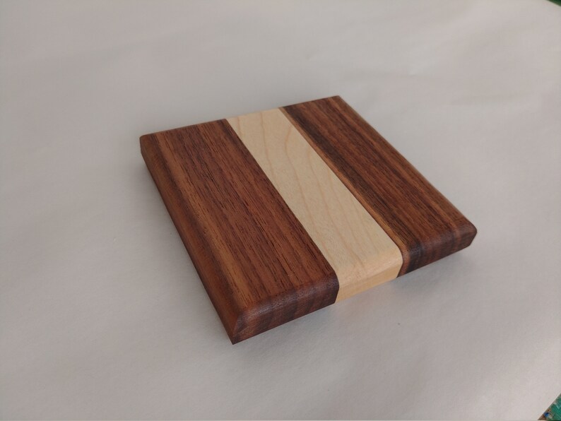May include: Square wooden cutting board with a dark brown walnut exterior and a light maple wood stripe down the center. The board has rounded edges and a smooth finish, suitable for kitchen use.