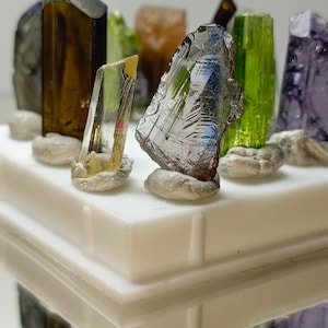 May include: A collection of various colorful crystals and gemstones, including green, brown, and purple hues. The crystals are displayed on a white platform, with some resting on small, gray stones. The crystals have a reflective surface.