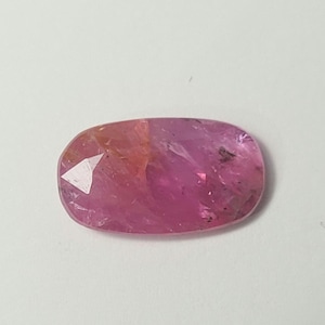 May include: An oval-shaped, faceted gemstone displaying a vibrant pink hue with subtle orange undertones. The stone features a polished surface with visible internal inclusions and light-reflecting facets across its translucent, elongated body.