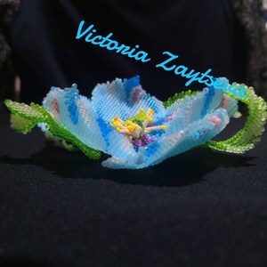 May include: A delicate blue flower crafted from beads, with green leaves and a colorful center. The flower is on a black background with the text "Victoria Zaytseva" in blue.