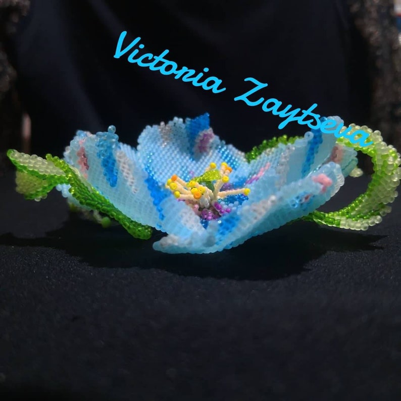 May include: A delicate blue flower crafted from beads, with green leaves and a colorful center. The flower is on a black background with the text "Victoria Zaytseva" in blue.