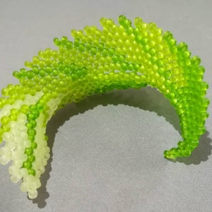 May include: A green and white beaded sculpture in the shape of a wave. The beads are arranged in a spiral pattern, creating a dynamic and textured effect.