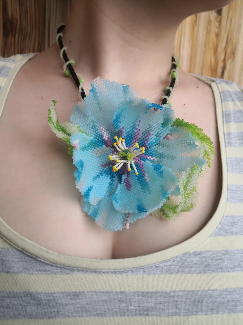 May include: A blue beaded flower necklace with green leaves and a black cord. The flower is made of blue, purple, and white beads.