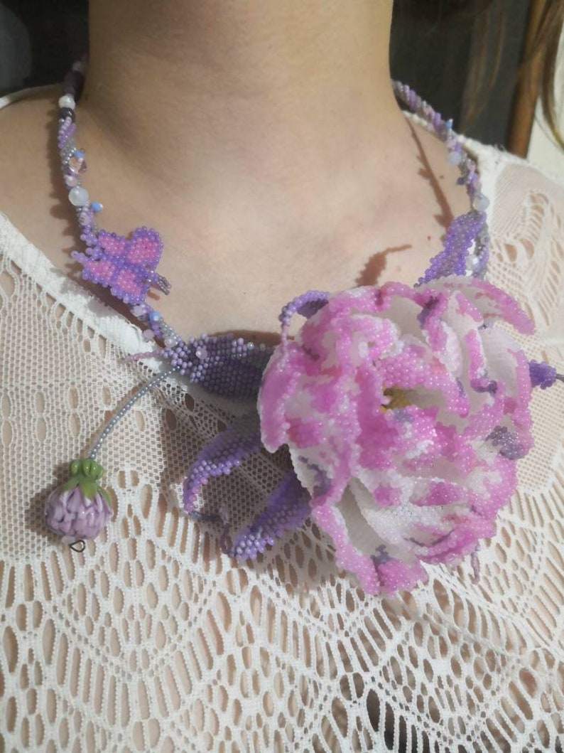 May include: A beaded necklace with a large pink flower and a smaller purple flower. The necklace is made of small beads in shades of purple, pink, and white.