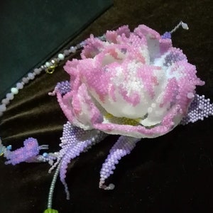 May include: A beaded flower necklace with a pink and white flower and purple leaves. The necklace is made of small beads and has a silver chain.