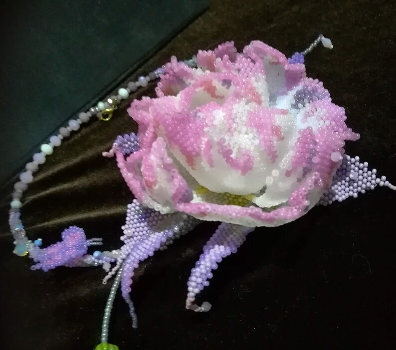May include: A beaded flower necklace with a pink and white flower and purple leaves. The necklace is made of small beads and has a silver chain.