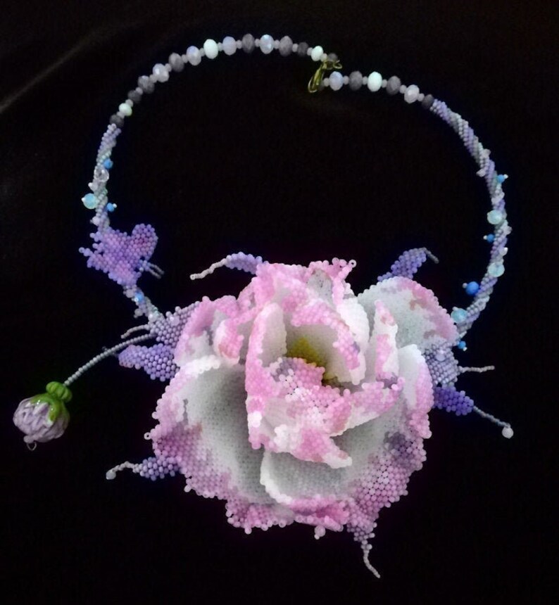May include: A delicate beaded necklace with a large, pink and white beaded flower pendant. The necklace is made of small, iridescent beads in shades of purple, blue, and white. The flower pendant is a realistic representation of a rose, with intricate details and a layered design.