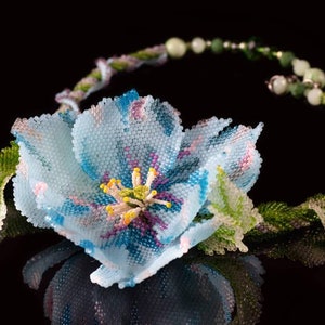 May include: A delicate beaded necklace featuring a light blue flower with pink and purple accents. The flower is surrounded by green leaves and the necklace is made of small beads.