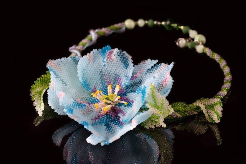 May include: A delicate beaded necklace featuring a light blue flower with pink and purple accents. The flower is surrounded by green leaves and the necklace is made of small beads.