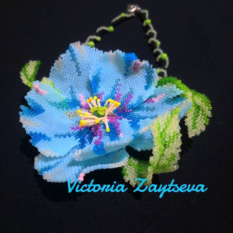 May include: A beaded flower necklace with a blue flower and green leaves. The flower has a yellow center with pink and purple accents. The necklace is made by Victoria Zaytseva.