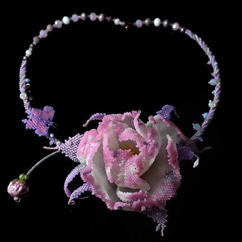 May include: A delicate beaded necklace with a large pink flower pendant. The flower is made of tiny beads and has a white center. The necklace is made of small, iridescent beads in shades of purple and blue.