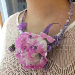May include: A delicate beaded necklace featuring a large pink and white flower with purple accents. The necklace is made of small beads and has a delicate, feminine look.