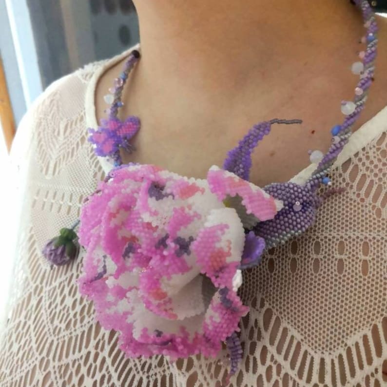 May include: A delicate beaded necklace featuring a large pink and white flower with purple accents. The necklace is made of small beads and has a delicate, feminine look.
