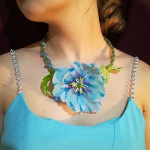 May include: A blue beaded flower necklace with green leaves and a silver chain. The necklace is worn by a person with a blue dress.