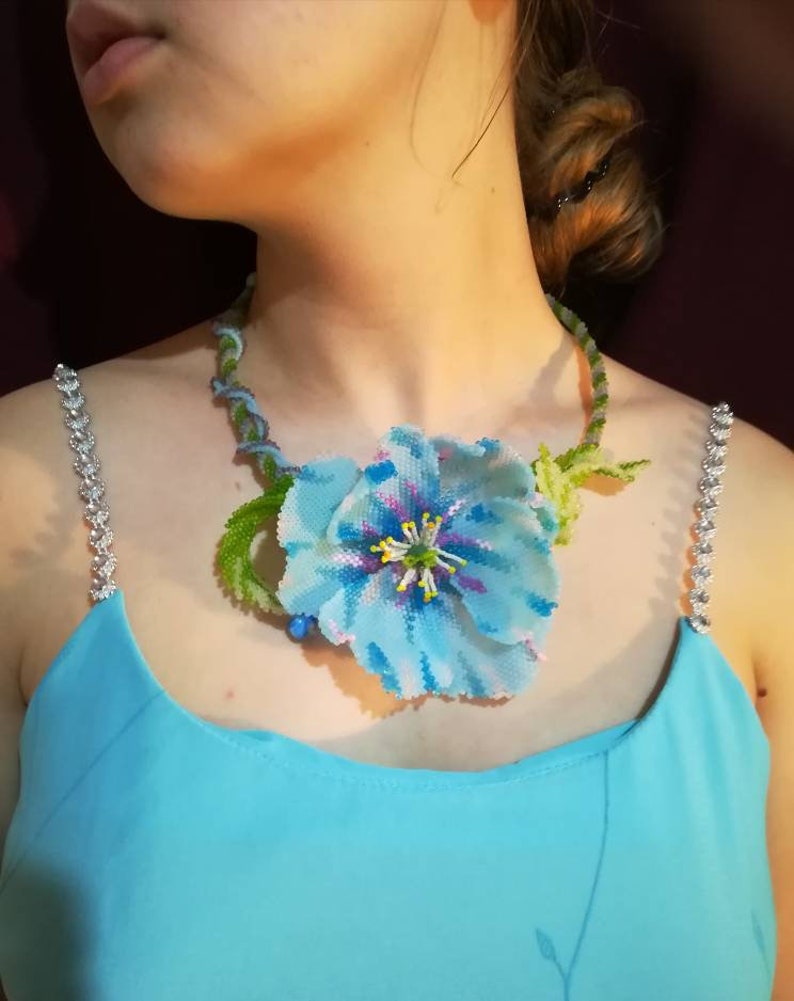 May include: A blue beaded flower necklace with green leaves and a silver chain. The necklace is worn by a person with a blue dress.