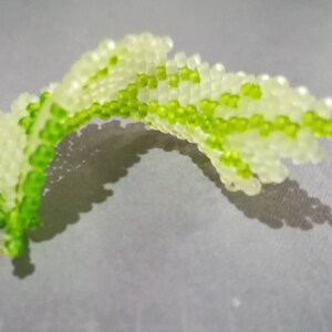 May include: A green and white beaded leaf, made of small round beads, is displayed on a gray surface.
