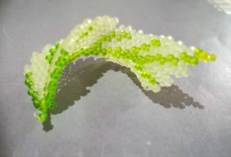 May include: A green and white beaded leaf, made of small round beads, is displayed on a gray surface.