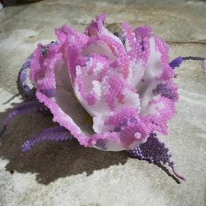 May include: A delicate, handmade flower crafted from purple and white beads. The flower has a layered, ruffled appearance and is a beautiful example of beadwork.