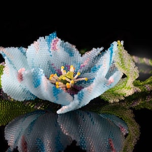 May include: A delicate blue flower crafted from beads, with a detailed center and green leaves. The flower is displayed on a black background.