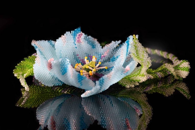 May include: A delicate blue flower crafted from beads, with a detailed center and green leaves. The flower is displayed on a black background.