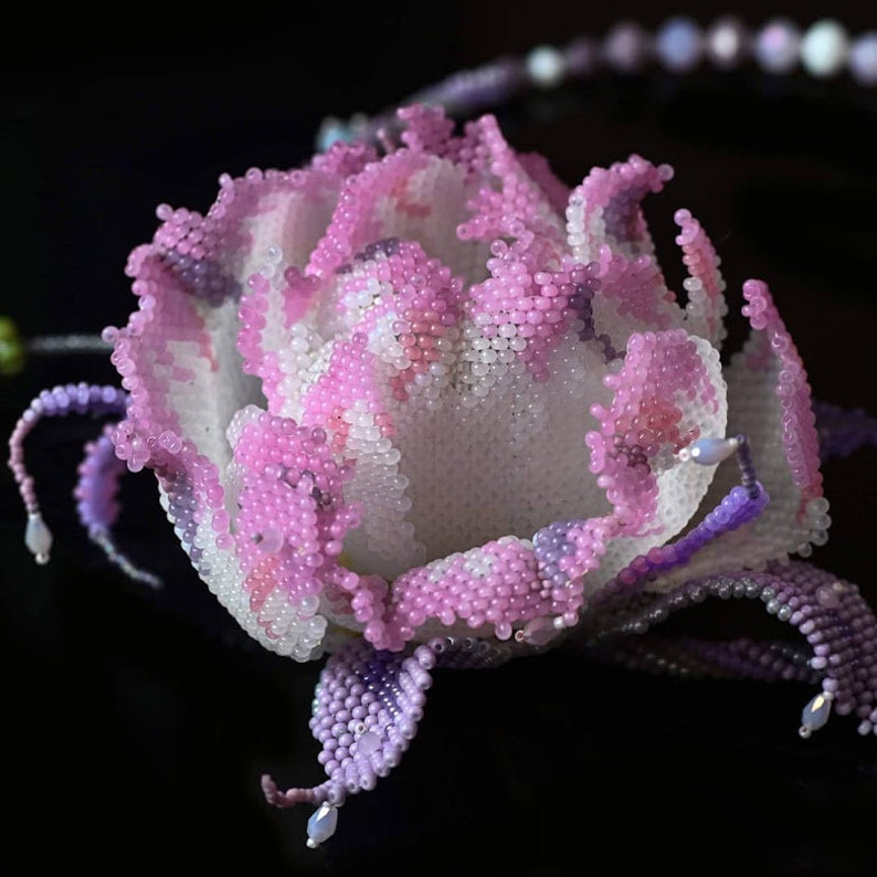 May include: A delicate pink and purple beaded flower, crafted with tiny beads, resembling a rose. The flower is detailed with intricate beadwork and has a realistic appearance.