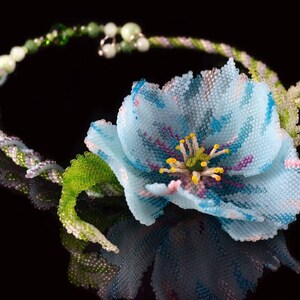 May include: A delicate beaded necklace with a light blue flower and green leaves. The flower has a pink and yellow center.