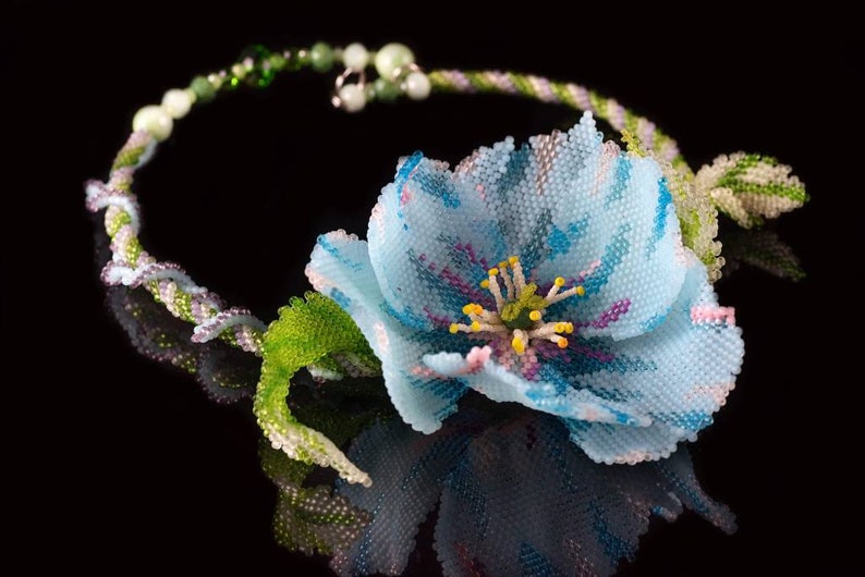 May include: A delicate beaded necklace with a light blue flower and green leaves. The flower has a pink and yellow center.