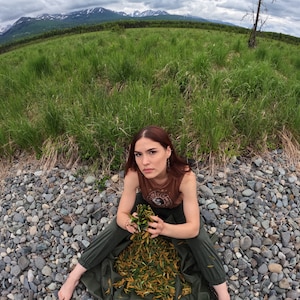 May include: A woman sits on a bed of small gray rocks, wearing a brown tank top and green pants. She is holding a handful of green leaves and looking directly at the camera.