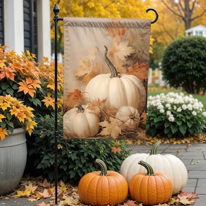 May include: A beige garden flag featuring a still-life of white pumpkins and autumn leaves. Two large white pumpkins and two smaller ones are depicted. The flag is displayed on a black metal stand, with orange pumpkins in the foreground.