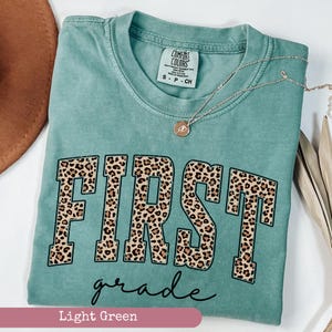 May include: Light green t-shirt with the word "FIRST" in large, black letters with a leopard print pattern. The word "grade" is written in cursive below the word "FIRST".