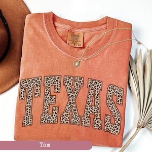 May include: A coral-colored t-shirt with the word "TEXAS" in large, leopard print letters. The shirt has a crew neck and short sleeves. A gold necklace with a pendant is draped across the shirt. The tag reads "Comfort Colors".