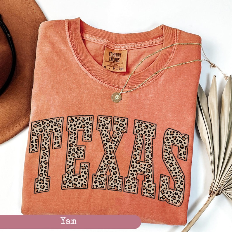 May include: A coral-colored t-shirt with the word "TEXAS" in large, leopard print letters. The shirt has a crew neck and short sleeves. A gold necklace with a pendant is draped across the shirt. The tag reads "Comfort Colors".