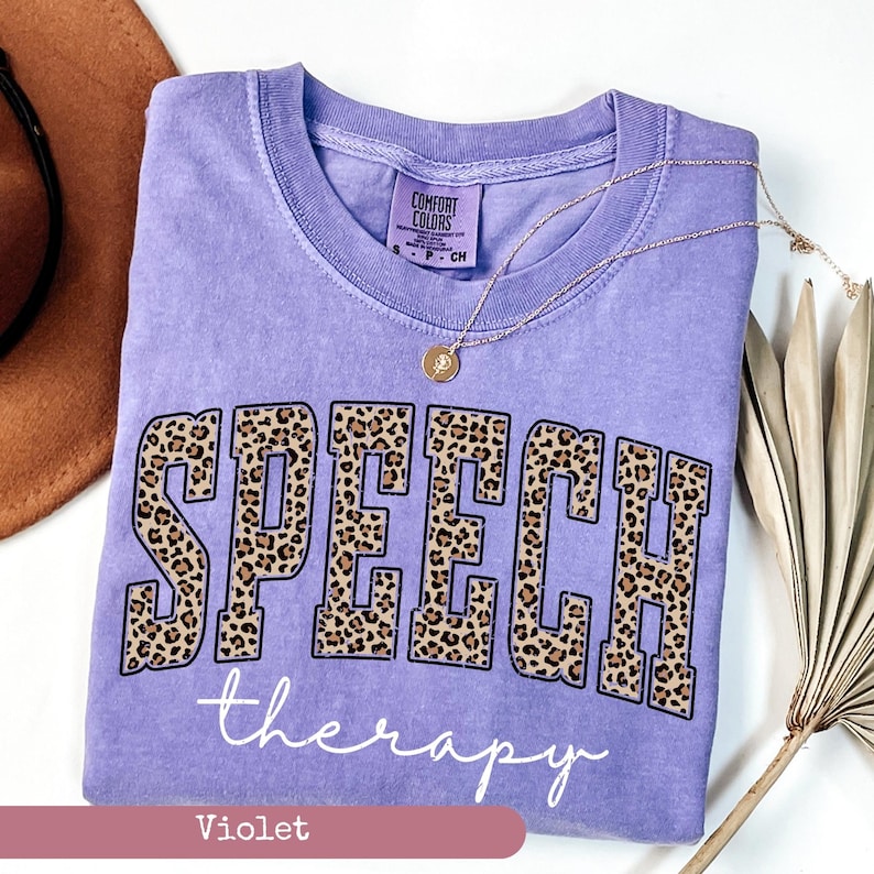 May include: A violet short-sleeved t-shirt with a crew neckline. The front features the word SPEECH in large block letters with a brown and black leopard print pattern, followed by the word therapy in white cursive script text.