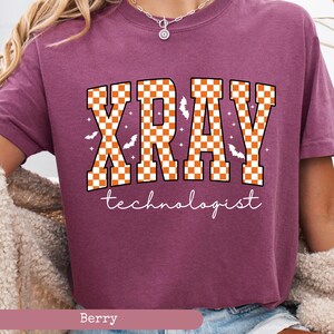 May include: Berry-colored t-shirt with the word "XRAY" in orange and white checkered letters. The word "technologist" is written in white script below the main text. The design includes bat and star graphics.
