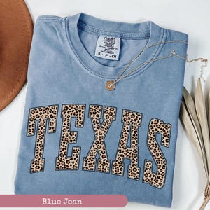 May include: A blue jean Comfort Colors t-shirt with the word "TEXAS" in large, leopard print letters. A gold necklace with a pendant rests on the shirt. The shirt is folded and has a small tag at the neckline.