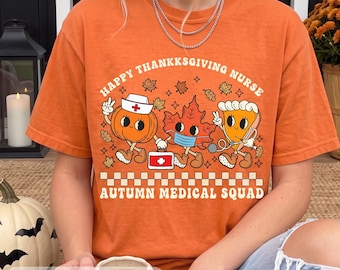 May include: An orange t-shirt featuring a graphic print with the text HAPPY THANKSGIVING NURSE and AUTUMN MEDICAL SQUAD. The design includes a pumpkin wearing a nurse cap, a maple leaf with a face mask, and a slice of pie with a stethoscope.