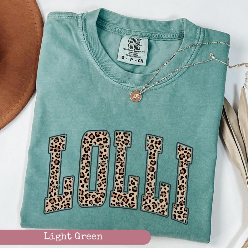 May include: A light green t-shirt with the word "LOLLI" printed in a leopard print pattern. The shirt is made of a soft, comfortable fabric and has a relaxed fit.