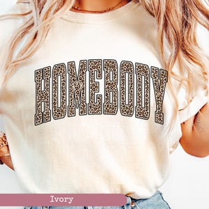 May include: Ivory-colored t-shirt with the word "HOMEBODY" in large, leopard print lettering across the front. The shirt has a crew neck and short sleeves. The bottom of the shirt is visible, and the color is labeled "Ivory".