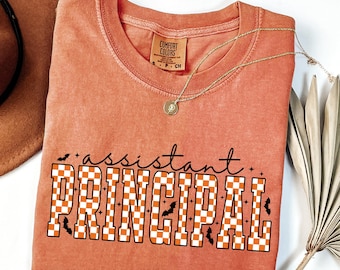 May include: A coral-colored t-shirt with the words "assistant PRINCIPAL" in a checkerboard pattern with bats. The shirt has a crew neck and a small tag that says "COMFORT COLORS". A gold necklace with a pendant is draped across the shirt.