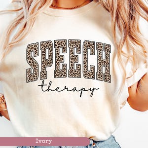 May include: An ivory t-shirt featuring the word SPEECH in large, block-style letters with a brown and black leopard print pattern. Below the word SPEECH, the word therapy is printed in a simple, black, lowercase cursive script font.