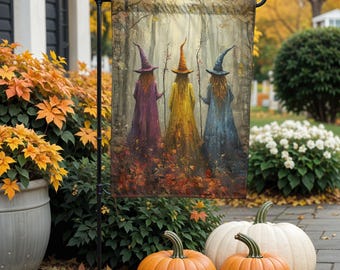 May include: A garden flag featuring three witches in colorful robes and pointed hats, walking through a forest with autumn leaves. The flag is displayed outdoors with pumpkins and fall foliage.