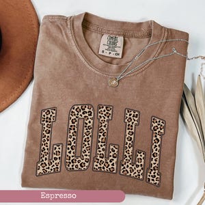 May include: Espresso brown t-shirt with the word "LOLLI" printed in large, bold letters. The letters are covered in a leopard print pattern.