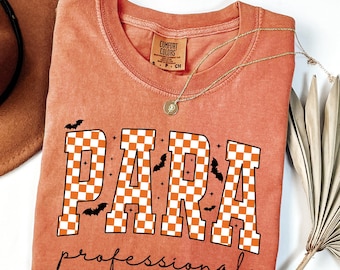 May include: Orange Comfort Colors t-shirt with the word "PARA" in large, checkered orange and white letters, and the word "professional" in a script font. Small black bats and stars are scattered around the word "PARA". A gold necklace with a pendant is draped on the shirt.