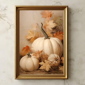 May include: A framed autumn still life featuring three white pumpkins of varying sizes, surrounded by orange and yellow fall leaves. The artwork is set against a soft, neutral background, and framed in a gold-colored frame.