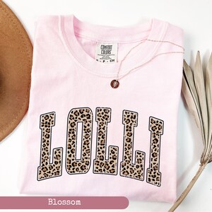 May include: Pink t-shirt with the word "LOLLI" printed in a leopard print font. The shirt is a light pink color and has a crew neck. The word "LOLLI" is in a large, bold font and is centered on the shirt.